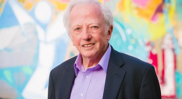 Hal Higdon, smiling, in a dark blazer and lavender shirt in front of a colorful mural.