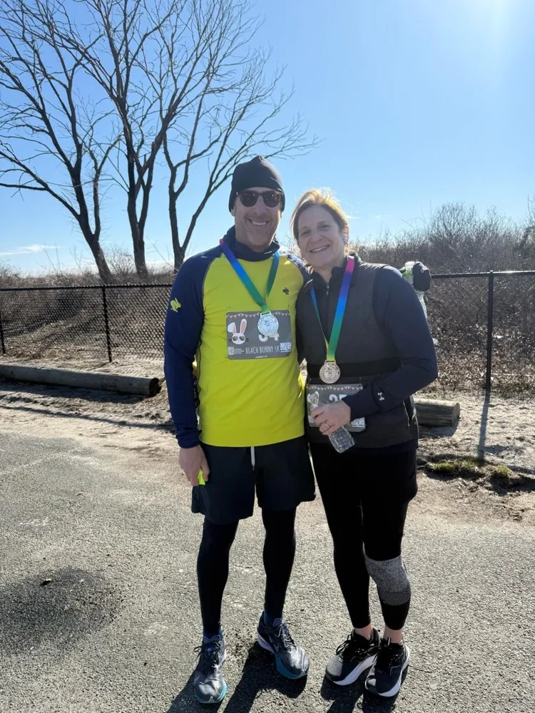 Two smiling runners wearing finisher medals in front of bare winter trees.