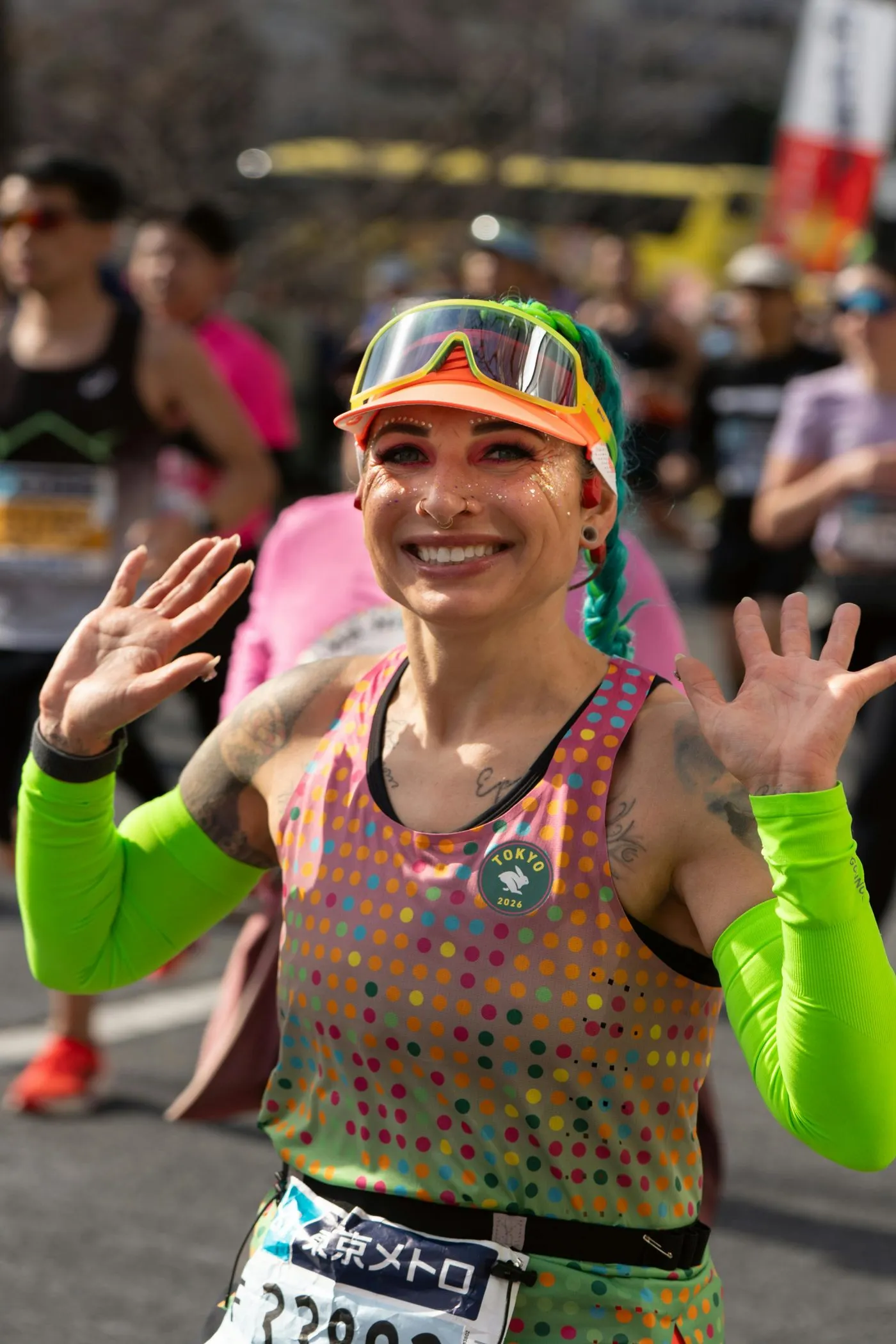 A smiling marathoner in a dotted singlet and bright green arm sleeves waves both hands mid-race.